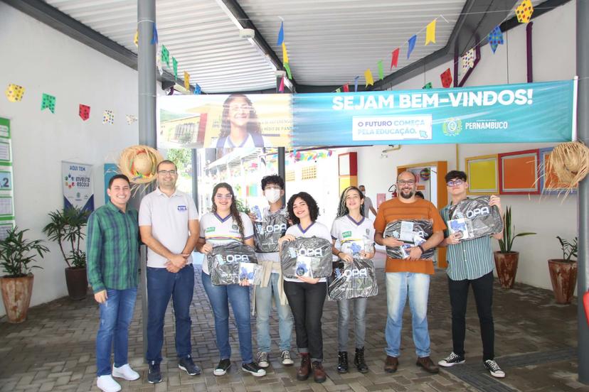 Grupo de estudantes e professores em escola decorada para festa, recebendo mochilas e kits escolares.  Banner de boas-vindas e decoração de bandeirinhas.