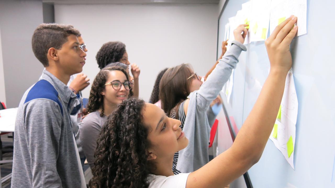 Um grupo de estudantes adolescentes colabora em um projeto, adicionando anotações coloridas a um quadro branco grande.