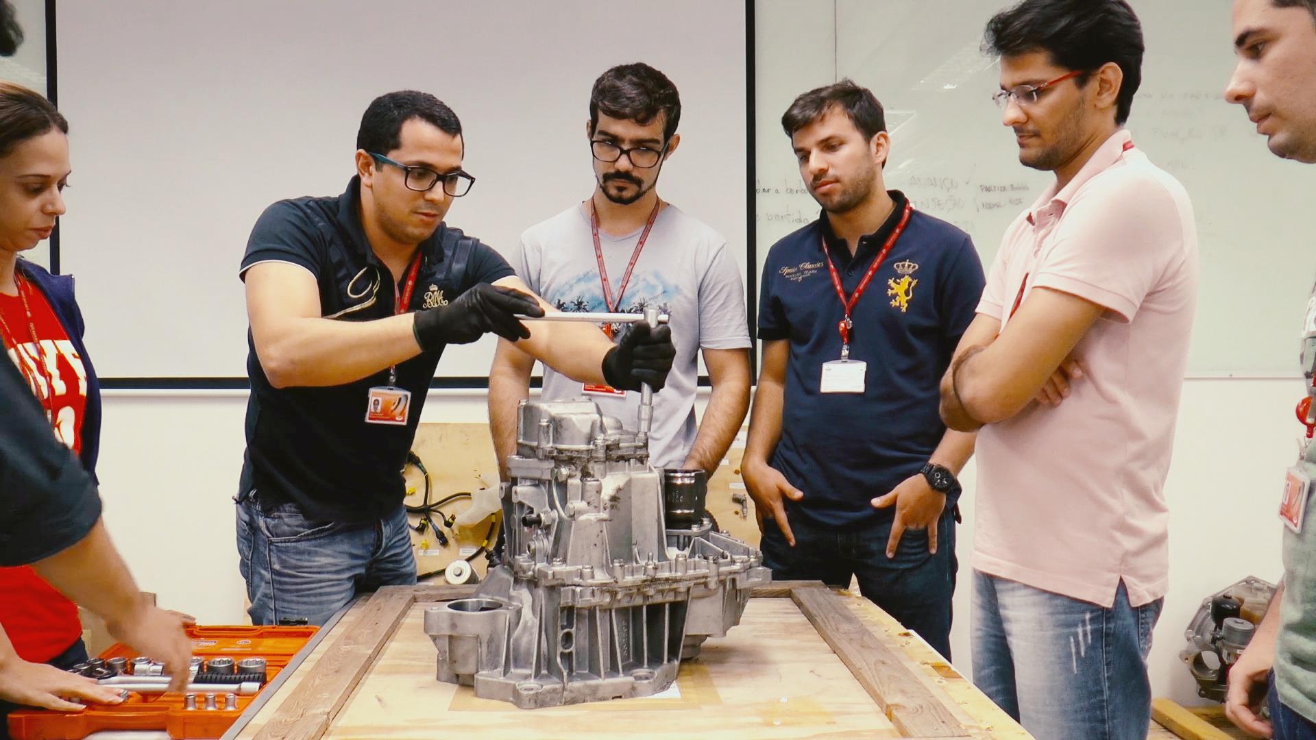 Um grupo de estudantes trabalha em conjunto na desmontagem de uma transmissão de carro, utilizando ferramentas manuais em uma mesa de madeira.