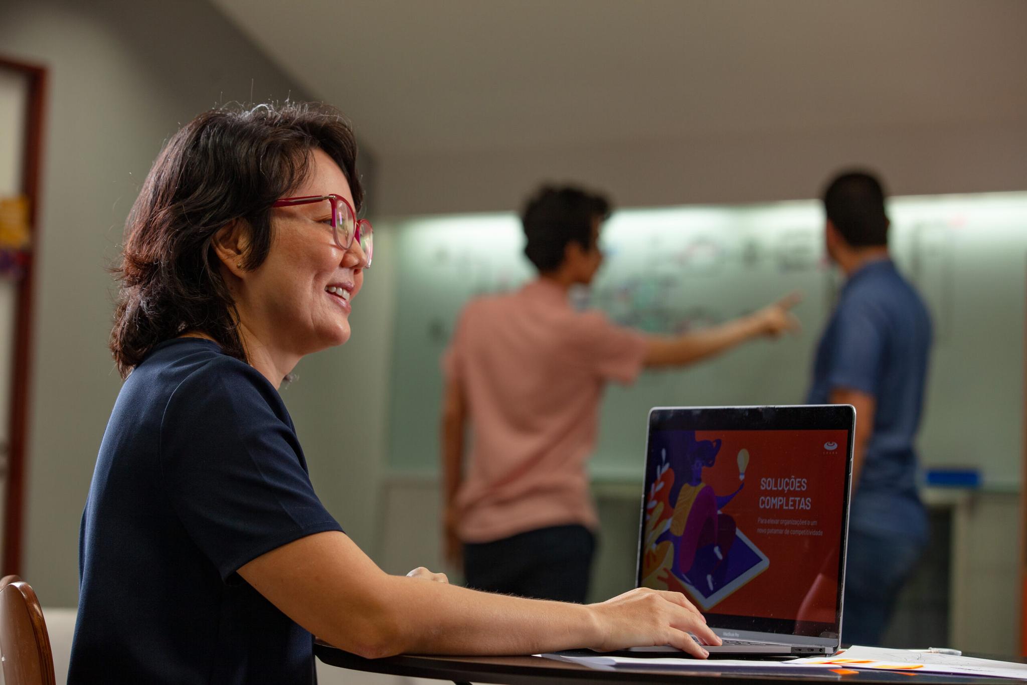 Uma mulher sorridente usando óculos senta-se a uma mesa com um laptop aberto mostrando um projeto, enquanto duas pessoas em segundo plano gesticulam para um quadro branco.