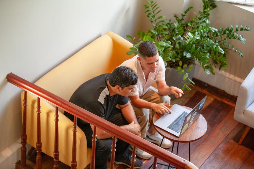 Dois homens sentados em um sofá amarelo, olhando para um laptop em uma mesa de madeira.  Um homem está apontando para a tela.