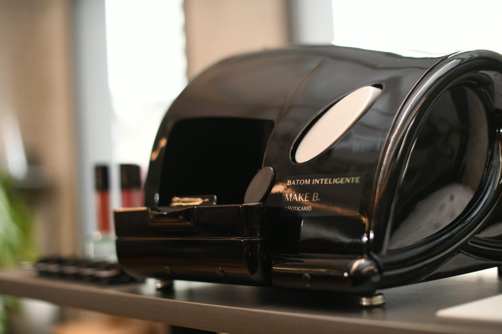 Close-up of a black Make B. intelligent lipstick machine by O Boticário, with some lipsticks blurred in the background.