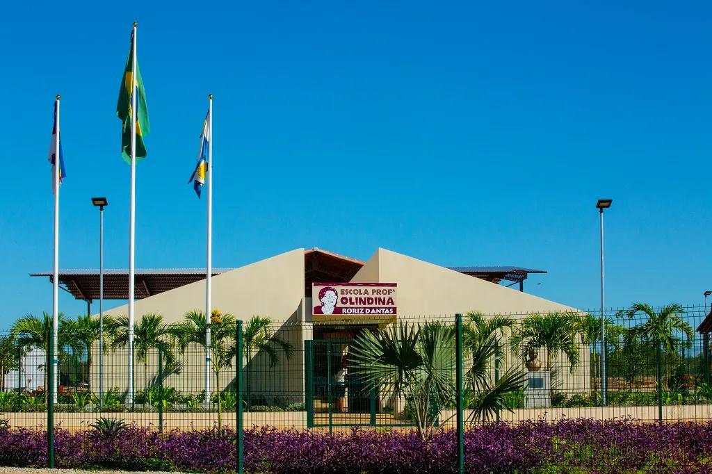 Fachada da Escola Olindina com três bandeiras à esquerda, palmeiras no jardim e céu azul ao fundo sob luz do dia.