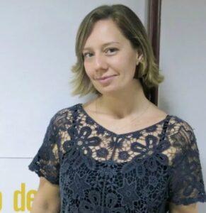 Uma mulher com cabelos na altura dos ombros, vestindo uma blusa de renda preta, está de pé contra um fundo simples, sorrindo gentilmente.