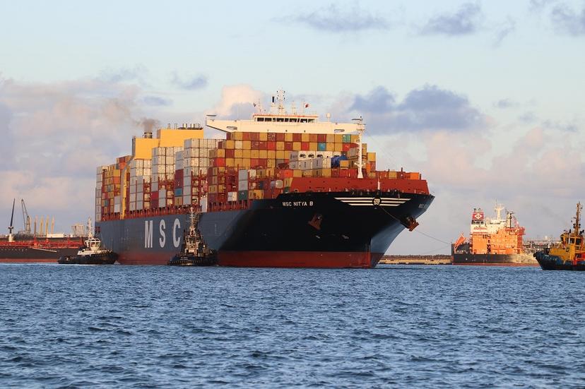 Um grande navio porta-contêineres MSC Nitya B no mar, cercado por rebocadores menores, com outros navios ao fundo.