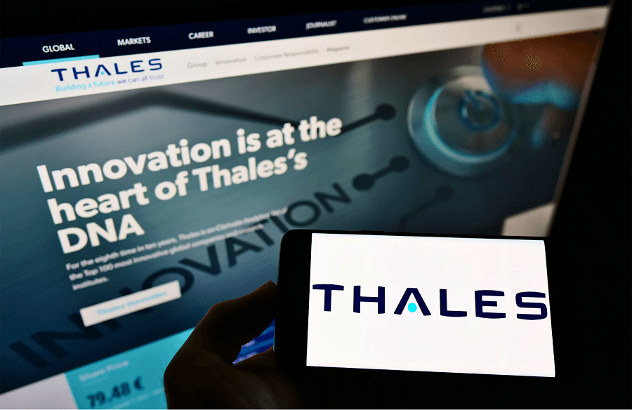 Um pessoa segurando um celular com a logo "Thales" e ao fundo uma tela de computador com o site deles aberto