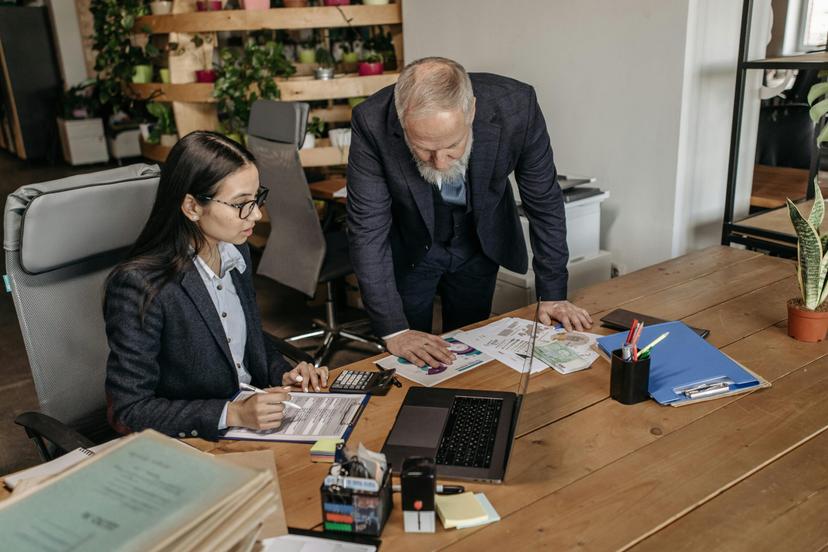 Um homem mais velho e uma mulher jovem, ambos em trajes de negócios, estão sentados a uma mesa de madeira, analisando documentos e usando um laptop.