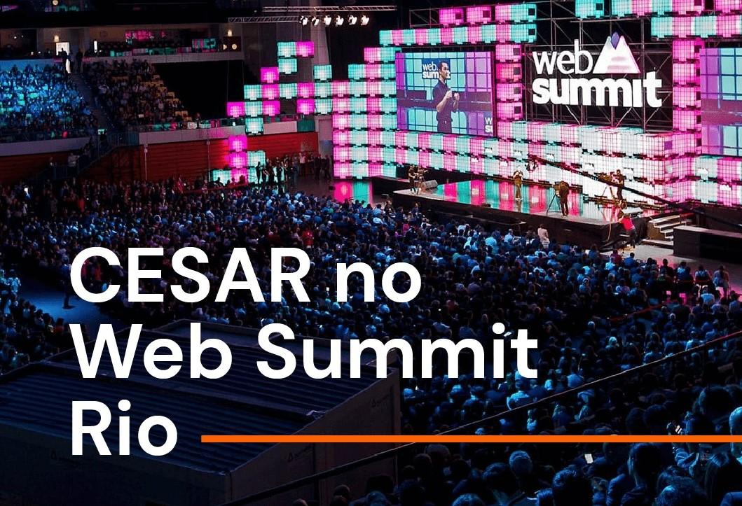 Grande plateia em um evento do Web Summit Rio, com um palco iluminado com o logo "Web Summit" exibido em grandes painéis de LED. Cesar está presente no evento.