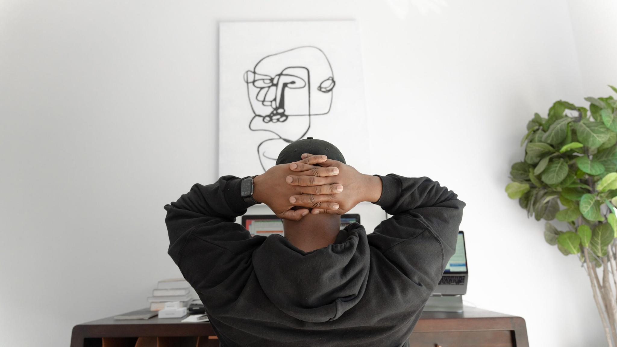 Home office setup: Person relaxing with hands behind head, facing away from the camera. Two laptops and abstract artwork are visible in the background.