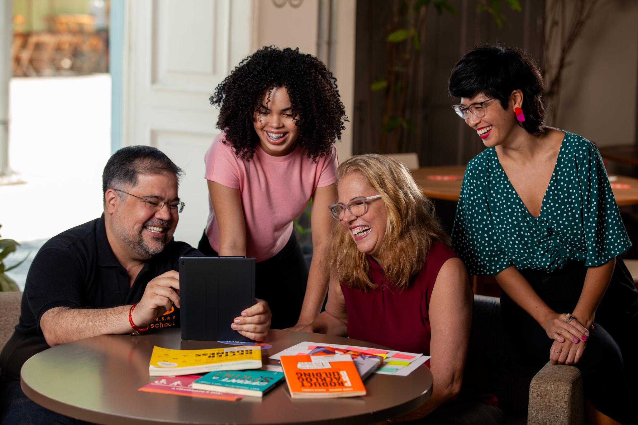 Quatro adultos sorrindo, reunidos em torno de um tablet e livros, parecem estar colaborando em um projeto ou estudando juntos.