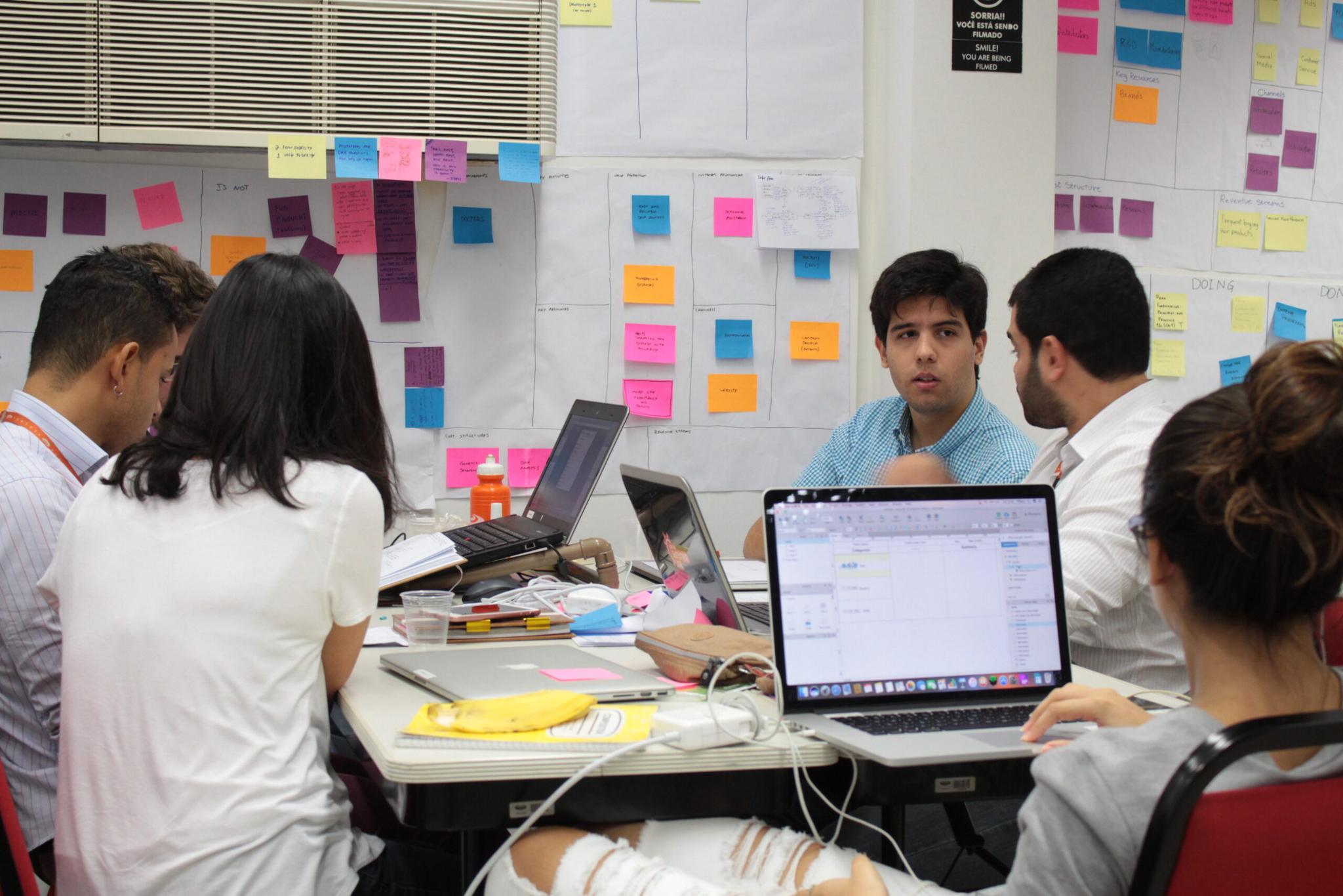 Um grupo de pessoas trabalha em computadores em uma sala de escritório com um quadro repleto de post-its coloridos.