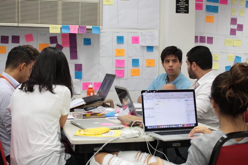 Um grupo de pessoas trabalha em computadores em uma sala de escritório com um quadro repleto de post-its coloridos.