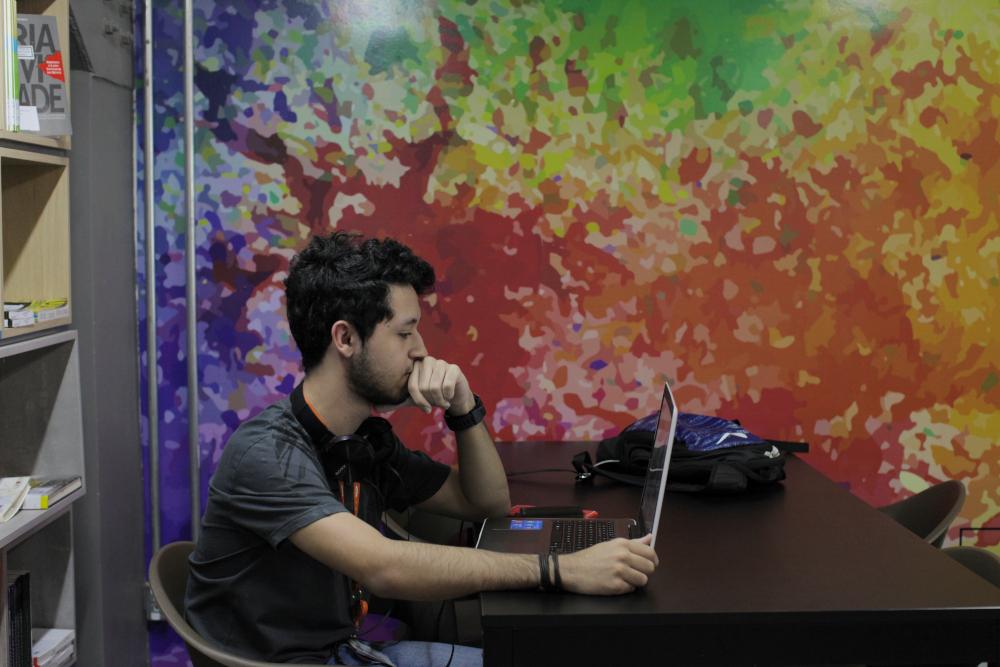 Homem com fone de ouvido usando laptop sentado a uma mesa, em frente a uma parede com pintura abstrata colorida.