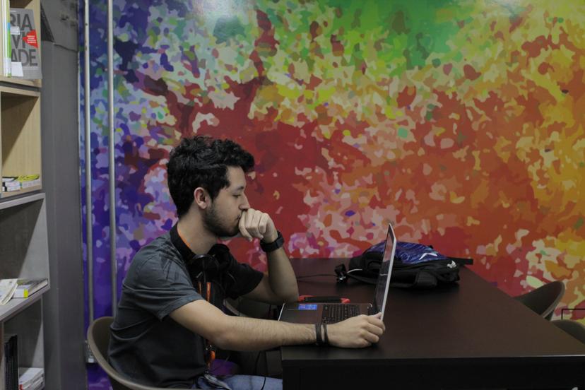 Homem com fone de ouvido usando laptop sentado a uma mesa, em frente a uma parede com pintura abstrata colorida.