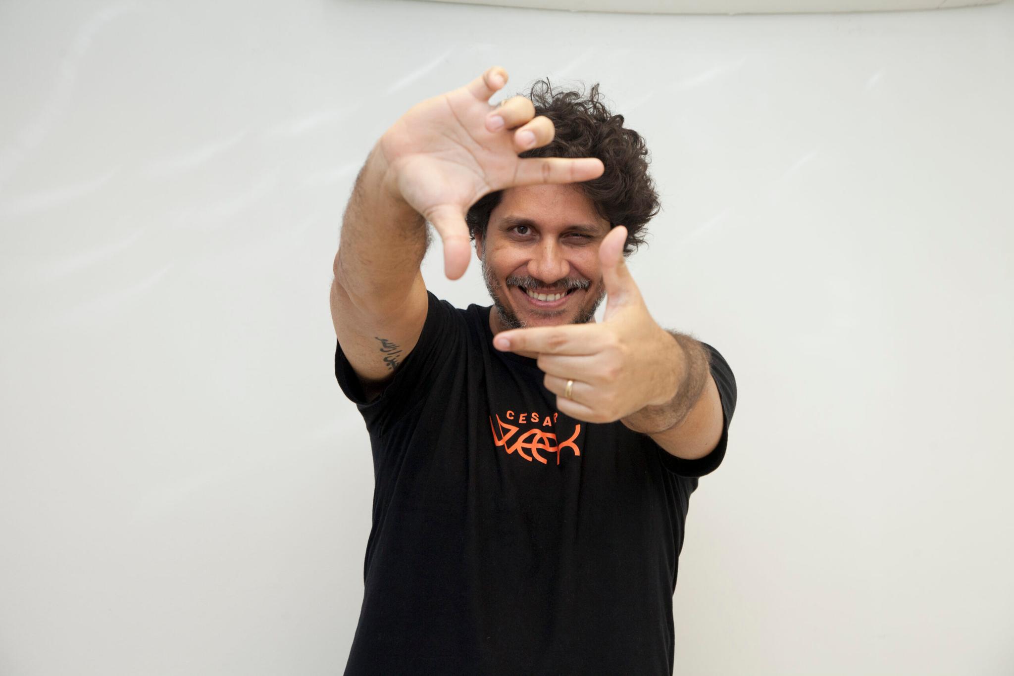 Homem sorridente com cabelos cacheados, usando camiseta preta, faz moldura com as mãos na frente do rosto contra fundo branco.