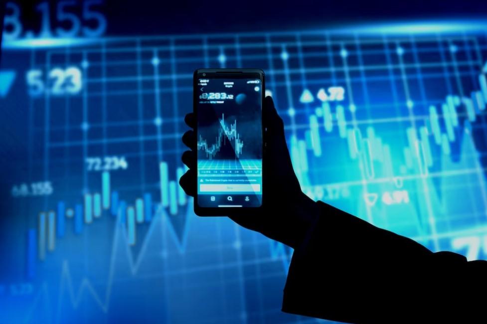 Silhueta de uma mão segurando um smartphone que exibe gráficos de ações em frente a um grande painel mostrando dados financeiros.