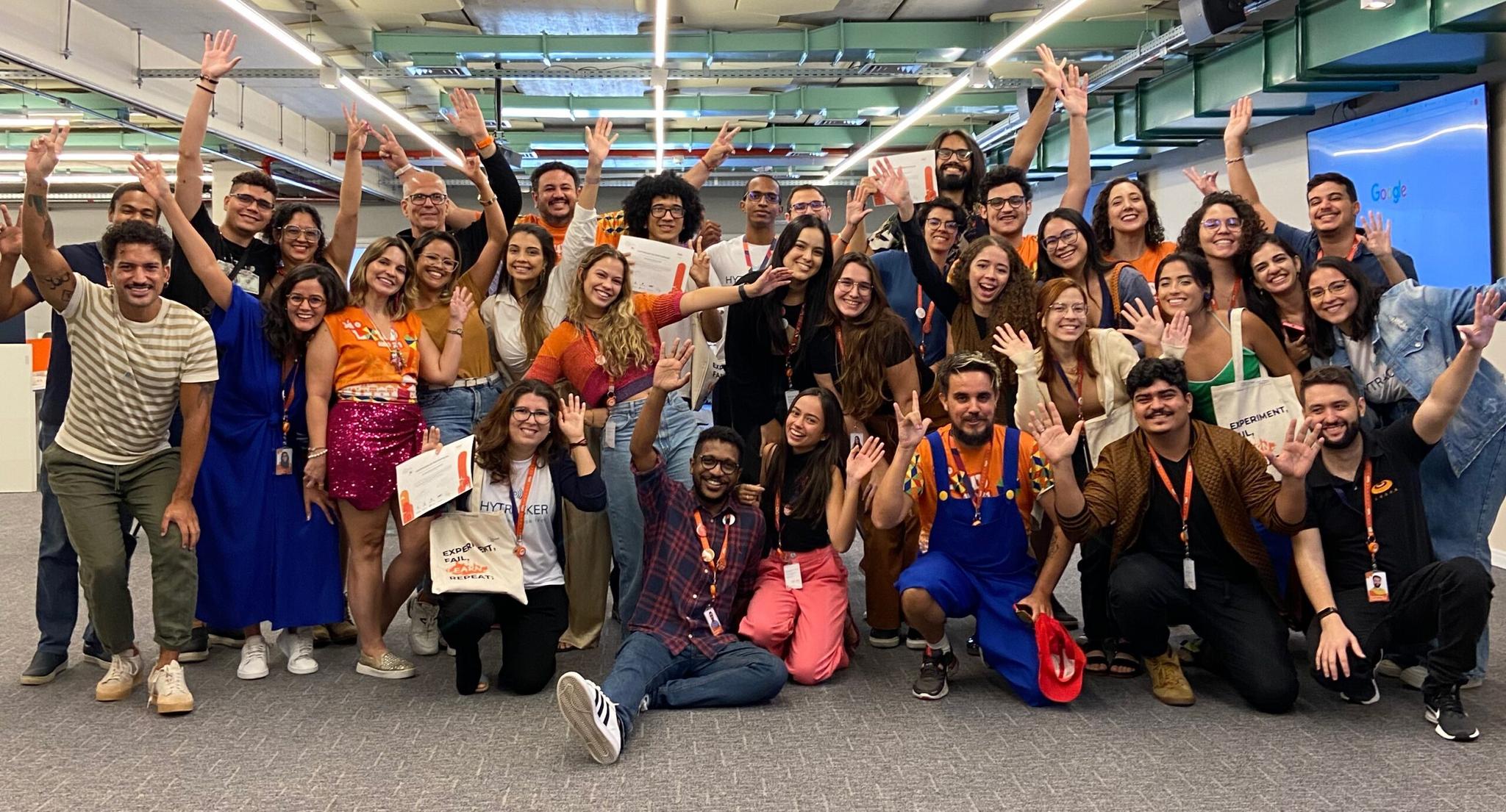 Grande grupo de pessoas sorrindo e com os braços para cima, em um ambiente de escritório.  Muitos estão vestindo roupas laranja e algumas pessoas usam fantasias.