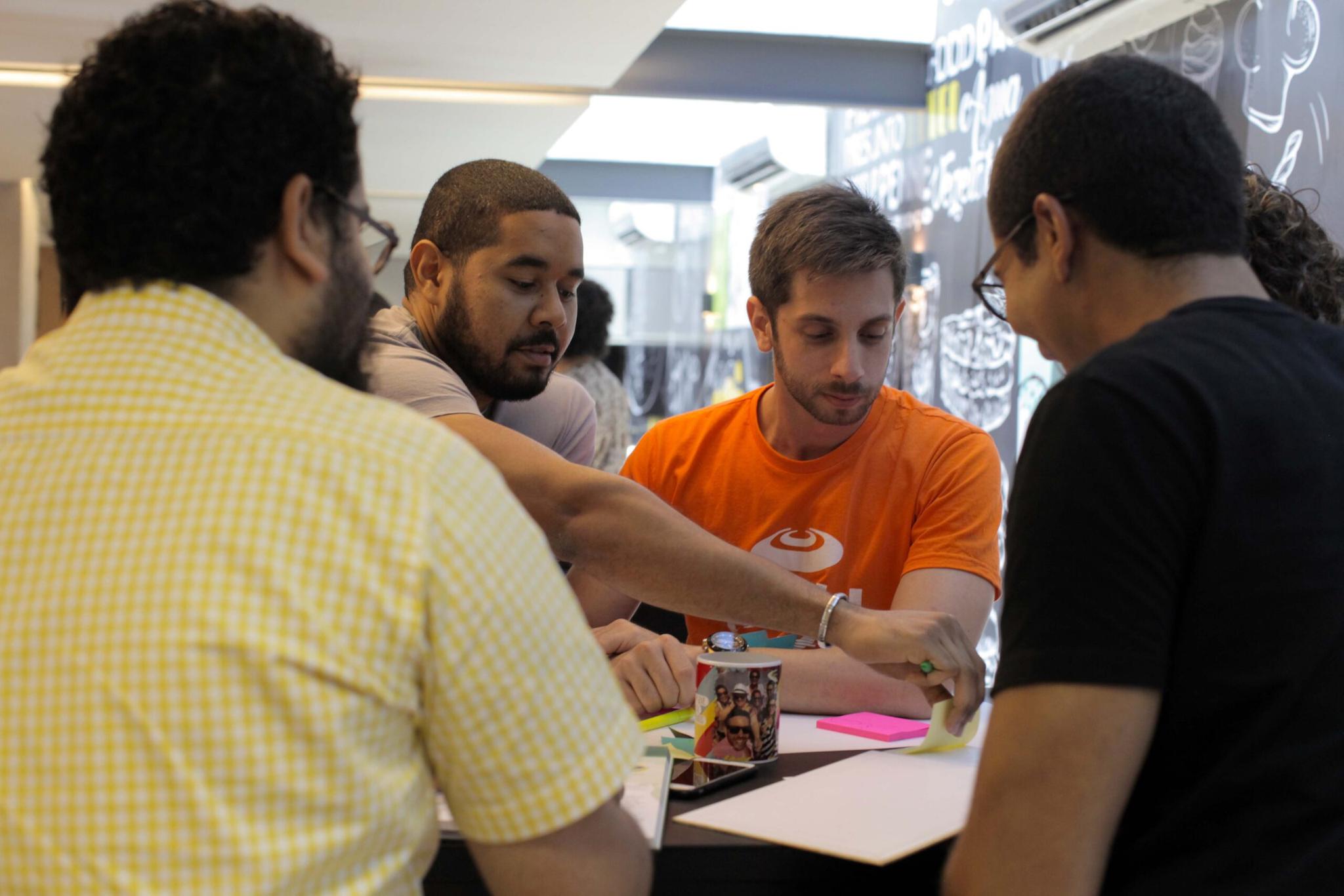 Quatro pessoas sentadas em torno de uma mesa, engajadas em uma atividade colaborativa. Elas parecem estar trabalhando em um projeto, utilizando post-its e uma caneca com fotos. A atmosfera é casual e produtiva.