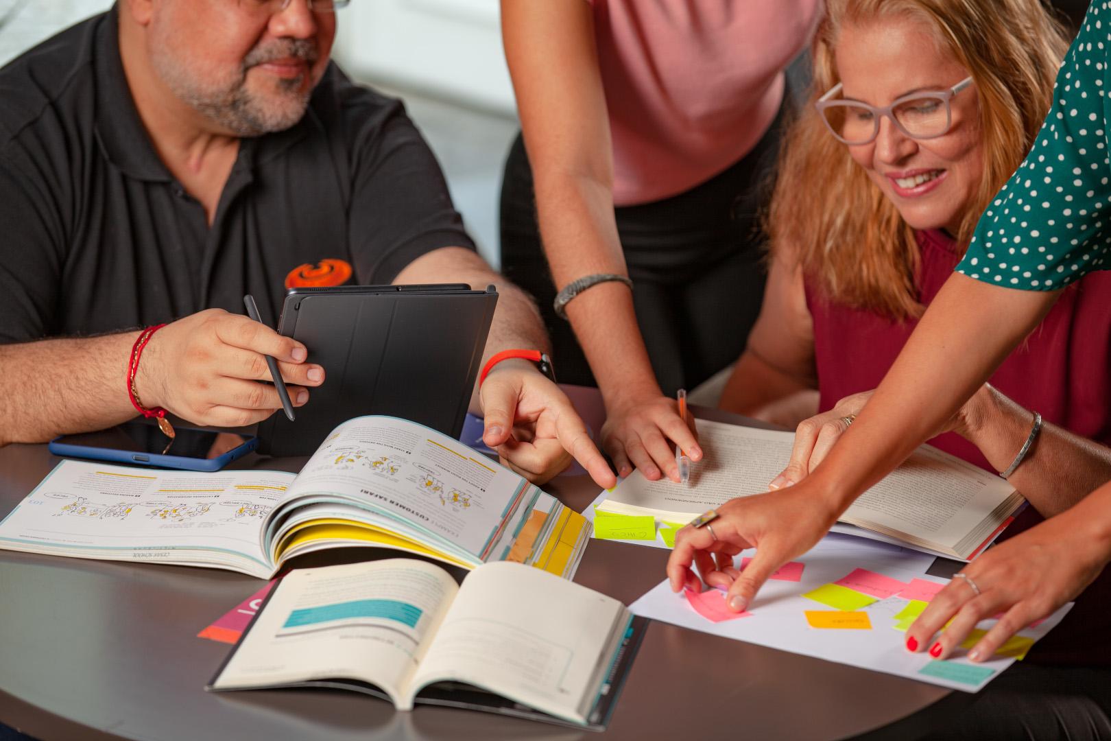 Um grupo de pessoas sentadas em torno de uma mesa, colaborando em um projeto com livros, cadernos, um tablet e notas adesivas.