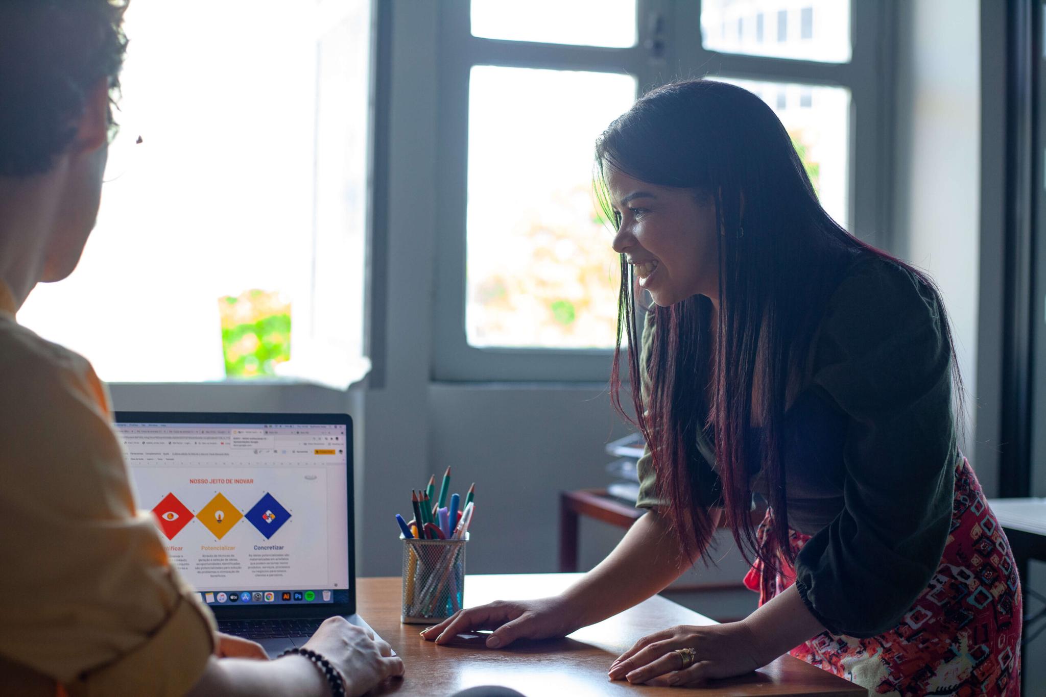 Duas pessoas sentadas à mesa, uma delas olhando para um laptop que exibe um gráfico, enquanto a outra se inclina sobre a mesa, conversando e interagindo.