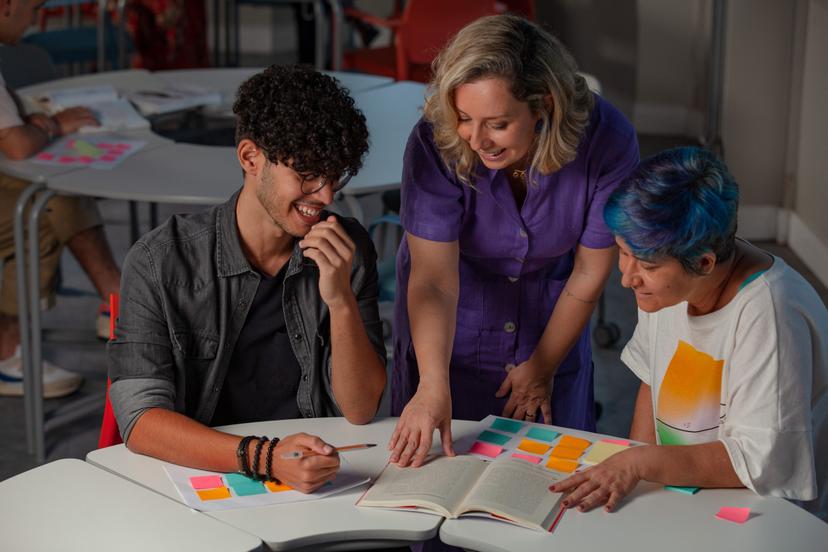Três pessoas, uma professora e dois estudantes, sentados à mesa, revisando um livro e anotações coloridas.