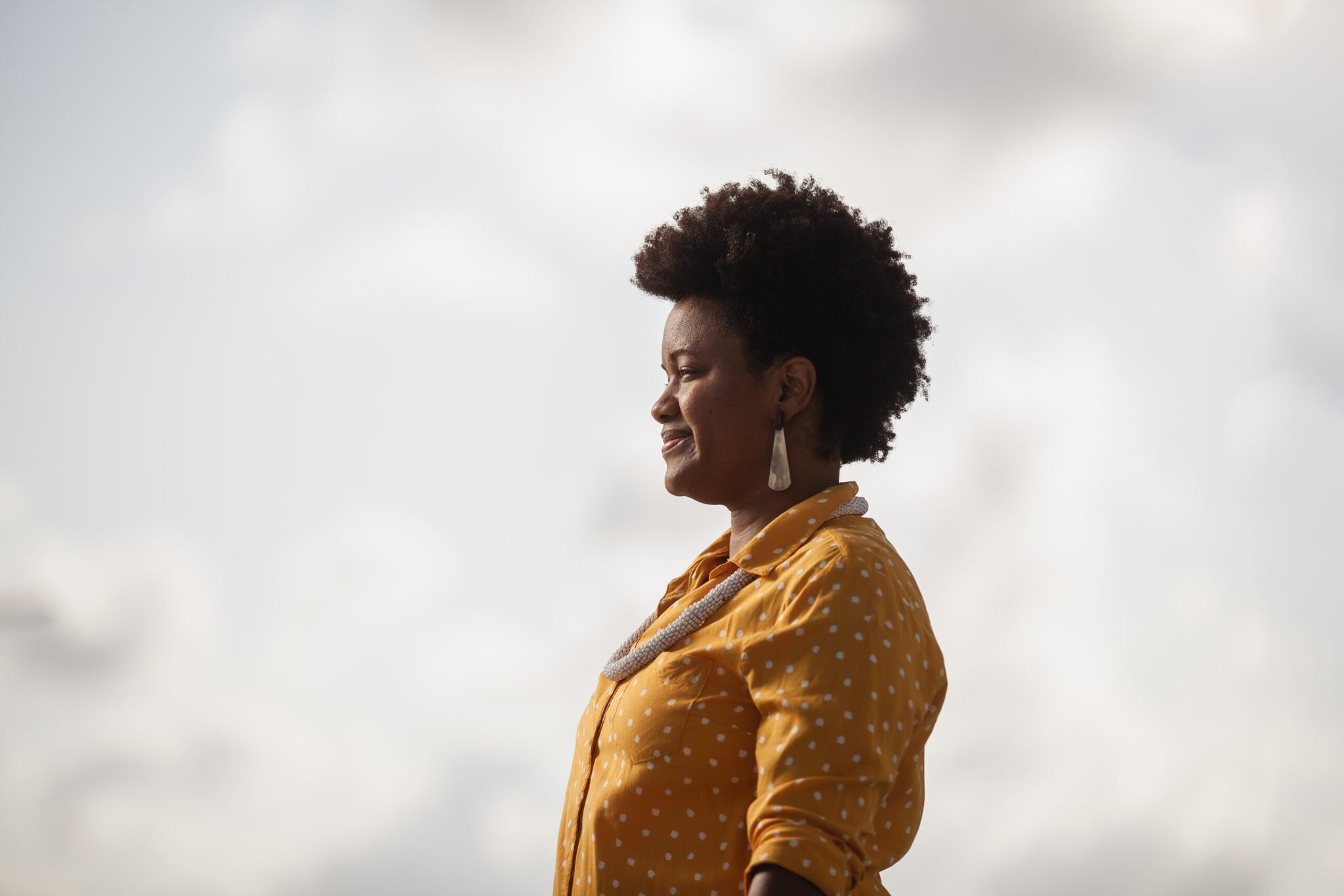 Uma mulher negra de cabelo curto e cacheado, usando um vestido amarelo com bolinhas e brincos, sorri enquanto olha para o horizonte nublado.