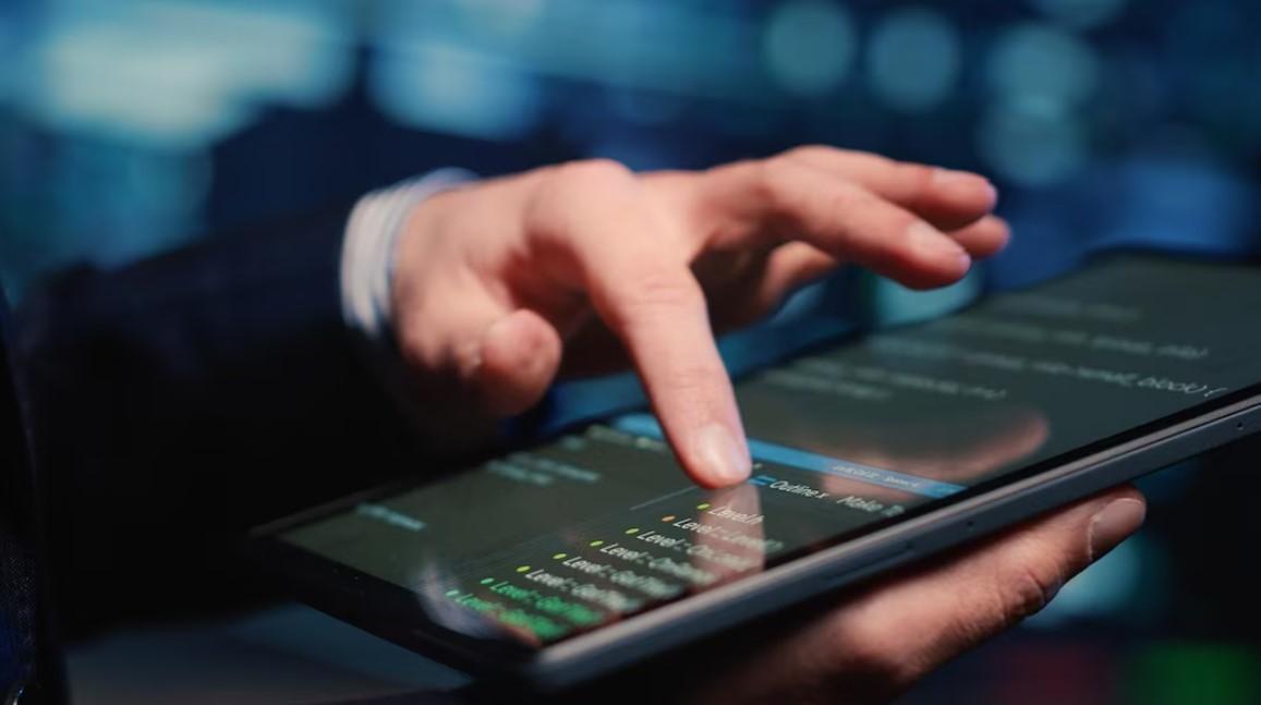 Close-up of a person's hand using a tablet displaying lines of code.  The background is blurred, emphasizing the technology.