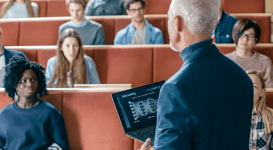 Professor de meia-idade dá aula sobre aprendizado profundo para uma turma de estudantes universitários em um auditório. Ele segura um laptop que exibe um diagrama de rede neural.