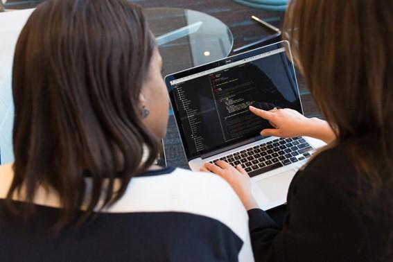 Duas mulheres sentadas juntas, olhando para um laptop que mostra código de programação na tela. Uma delas está apontando para a tela enquanto a outra digita.