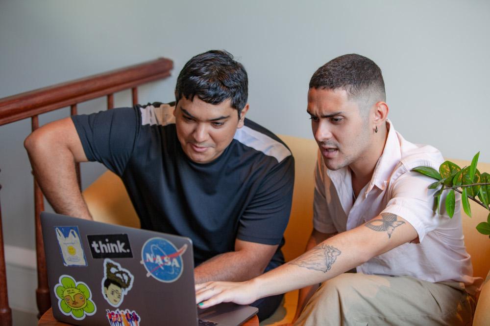 Dois homens sentados em um sofá, olhando para um laptop juntos. Um deles tem um braço tatuado e está apontando para a tela. O laptop tem vários adesivos.