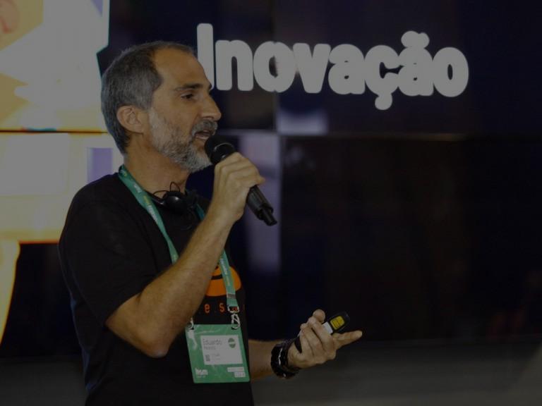 Homem de meia idade falando em um microfone em um evento com o letreiro "Inovação" ao fundo.