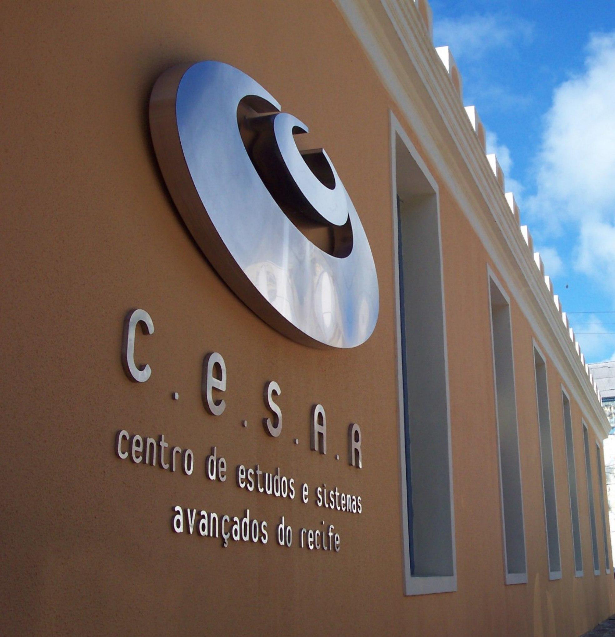 Logotipo metálico de C.E.S.A.R., Centro de Estudos e Sistemas Avançados do Recife, em uma parede de cor bege com várias janelas.
