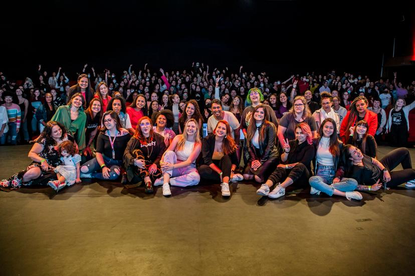 Um grande grupo de mulheres sentadas no chão, sorrindo para a câmera, na frente de uma multidão maior de pessoas em pé atrás delas.