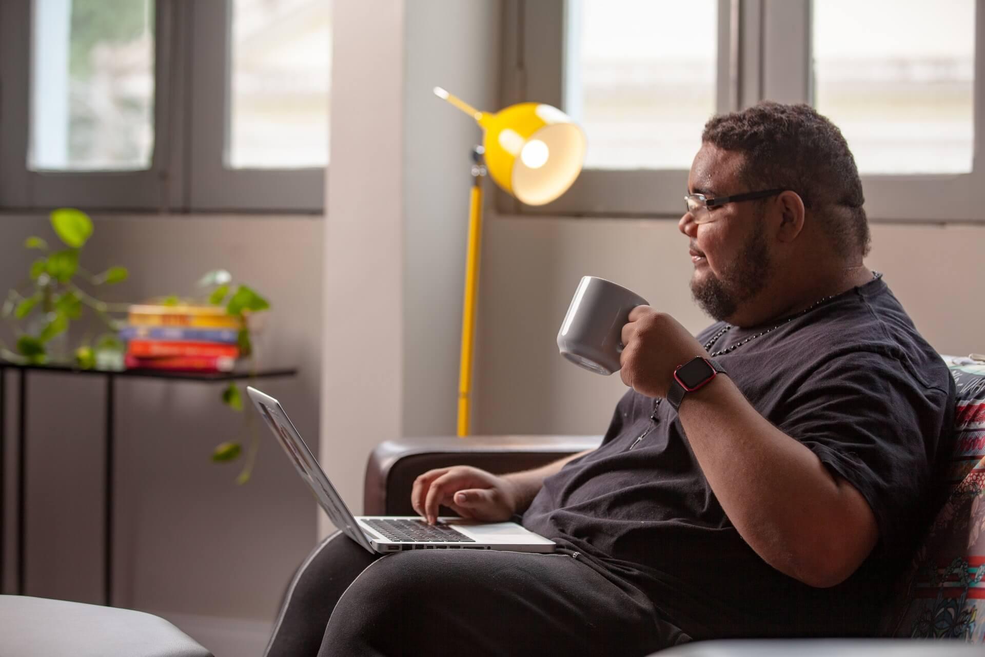 Homem com óculos e relógio sentado em um sofá, usando um laptop e bebendo café.