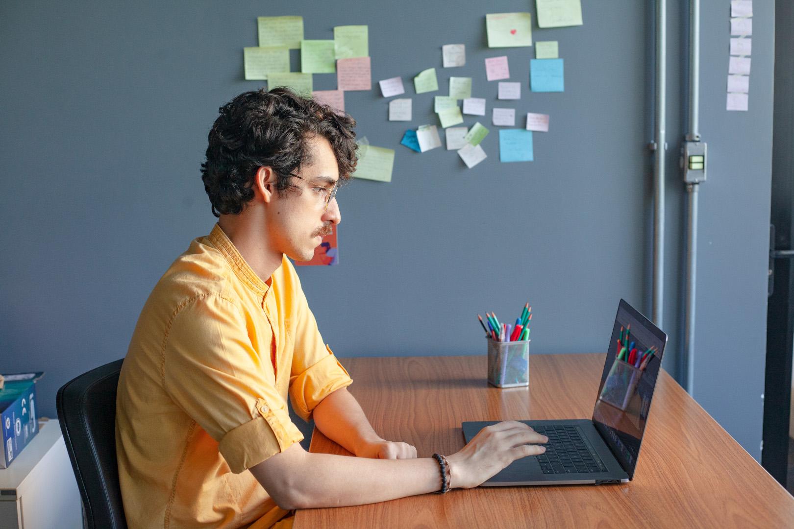 Homem de camisa amarela sentado à mesa, usando um laptop.  Há uma parede com post-its coloridos ao fundo.