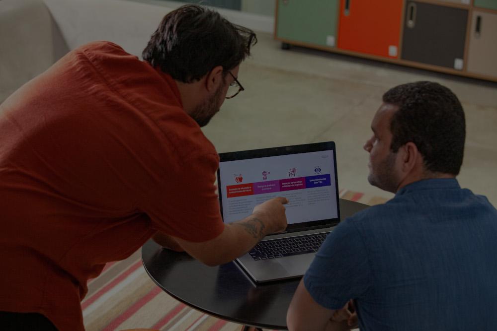 Dois homens sentados à mesa, um apontando para a tela de um laptop que mostra um gráfico.