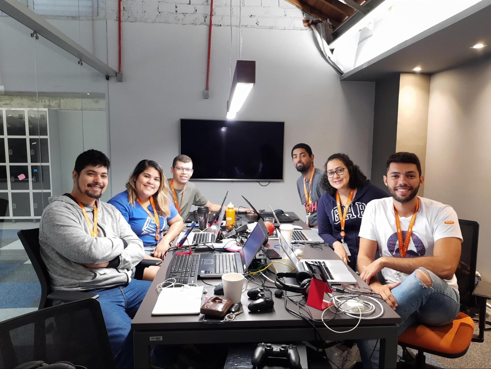 Sete jovens adultos sorrindo sentados em torno de uma mesa de escritório, com laptops e equipamentos eletrônicos.