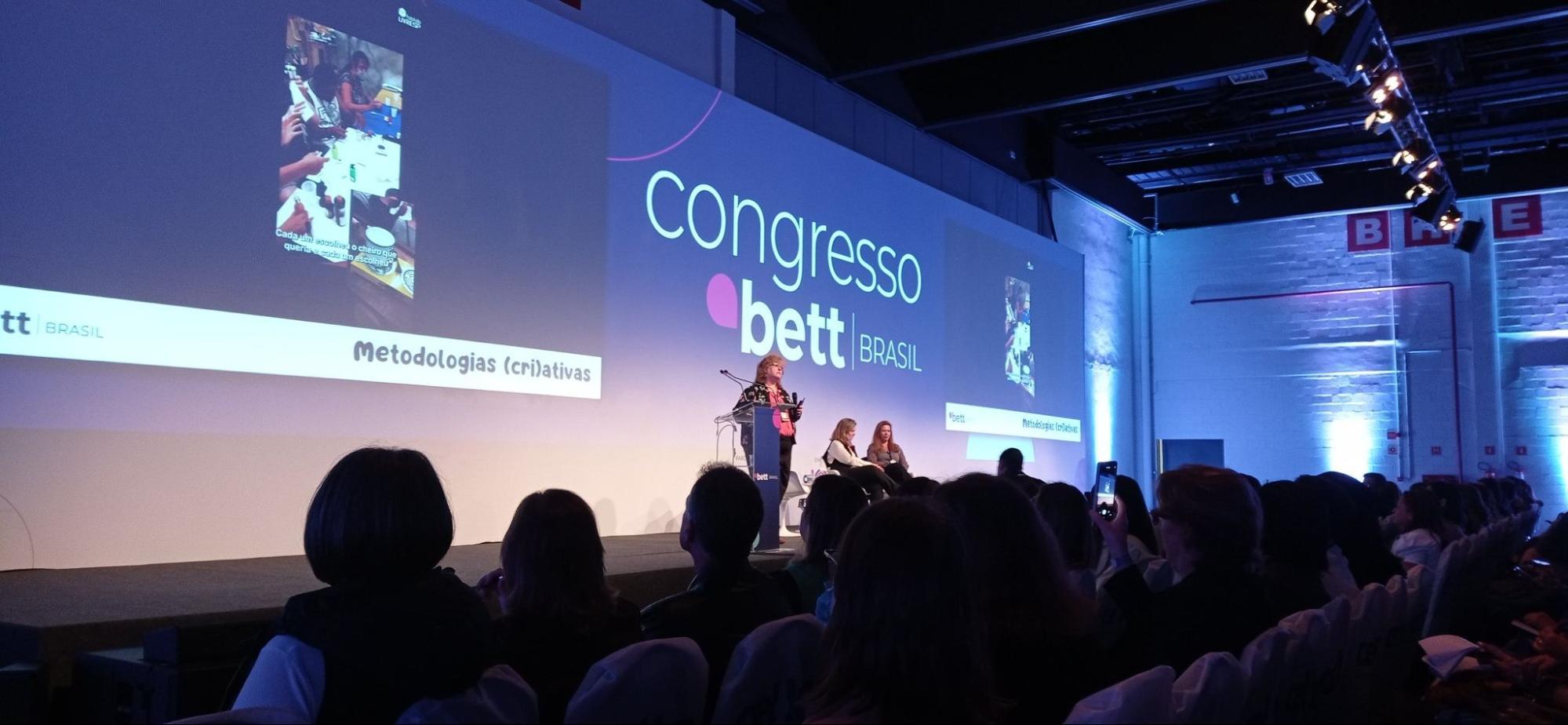 Uma palestrante em um palco, em um grande evento, com a logo do Congresso Bett Brasil exibida em um telão. A plateia está sentada e atenta.  Imagens adicionais são projetadas no telão.