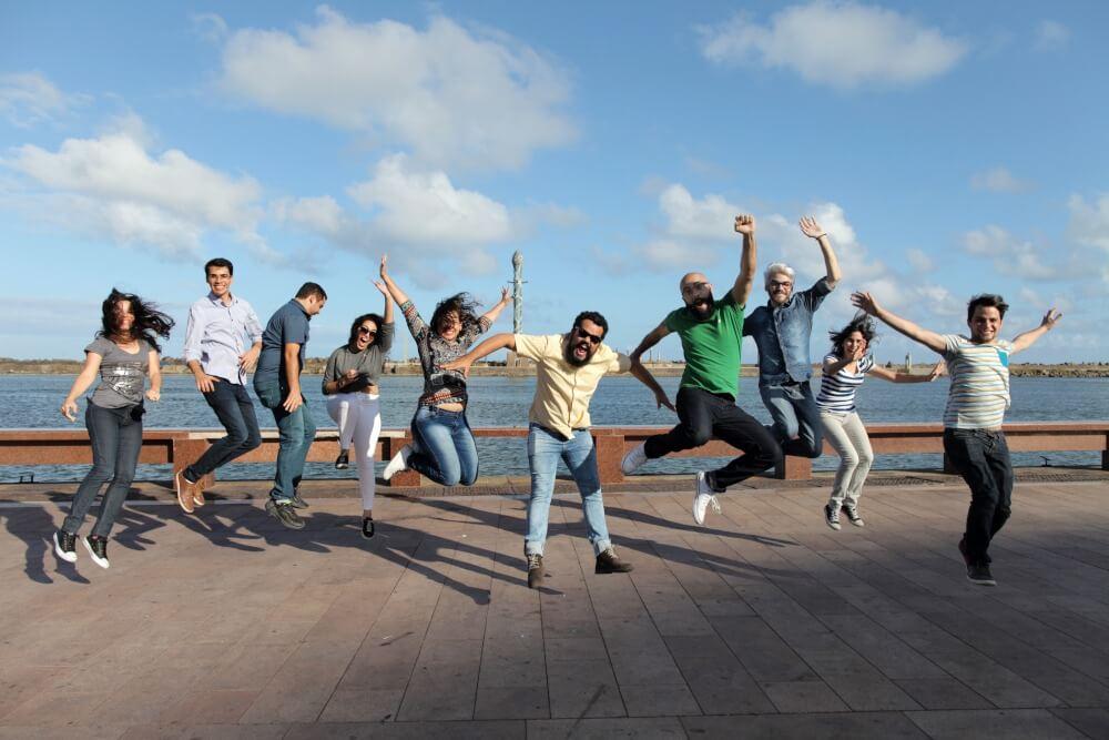 Um grupo de pessoas alegres salta no ar, numa esplanada junto ao mar, sob um céu azul e ensolarado.