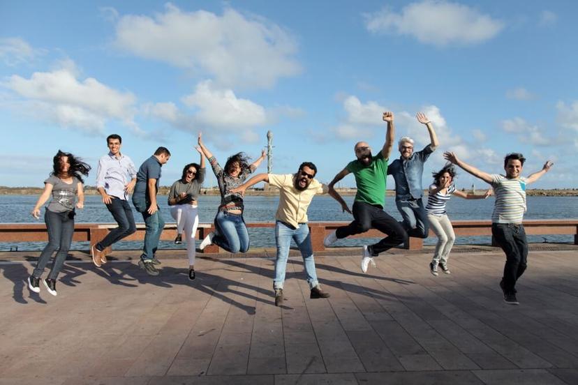 Um grupo de pessoas alegres salta no ar, numa esplanada junto ao mar, sob um céu azul e ensolarado.