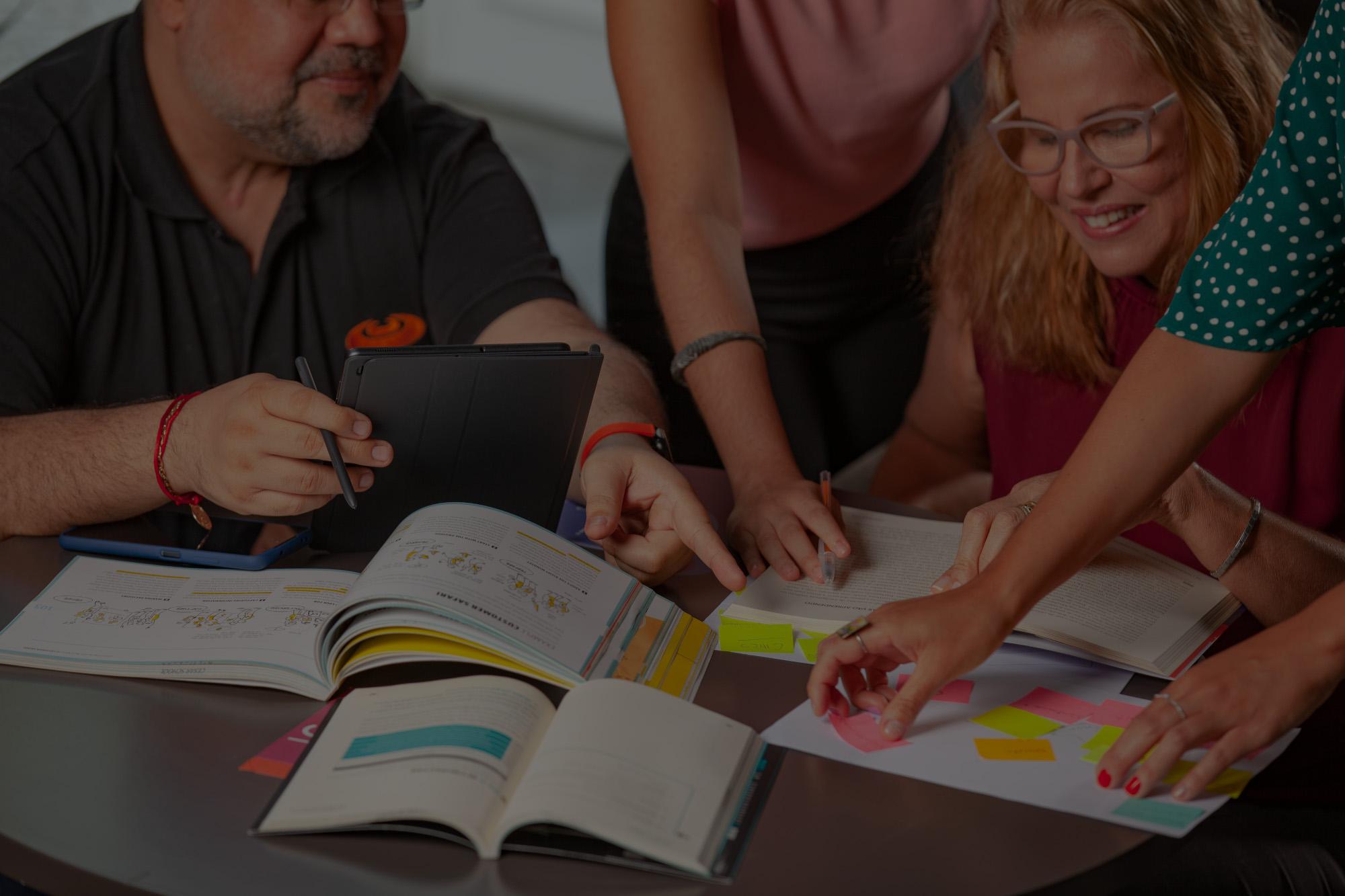 Um grupo de pessoas sentadas em torno de uma mesa, colaborando em um projeto. Livros, cadernos, um tablet e notas adesivas coloridas estão espalhados sobre a mesa. As pessoas estão envolvidas ativamente na discussão e no trabalho em equipe.