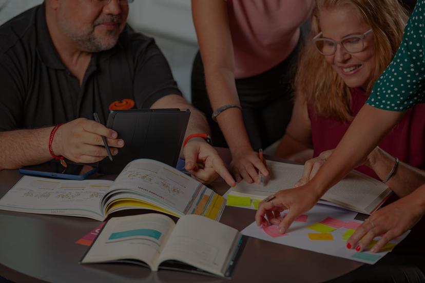 Um grupo de pessoas sentadas em torno de uma mesa, colaborando em um projeto. Livros, cadernos, um tablet e notas adesivas coloridas estão espalhados sobre a mesa. As pessoas estão envolvidas ativamente na discussão e no trabalho em equipe.