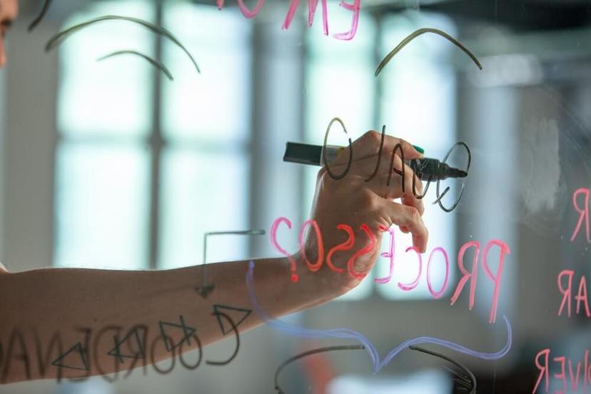 Um braço escreve "processo" e outros rabiscos em uma superfície de vidro transparente com canetas coloridas. A imagem está levemente desfocada, e pode-se ver uma janela ao fundo.