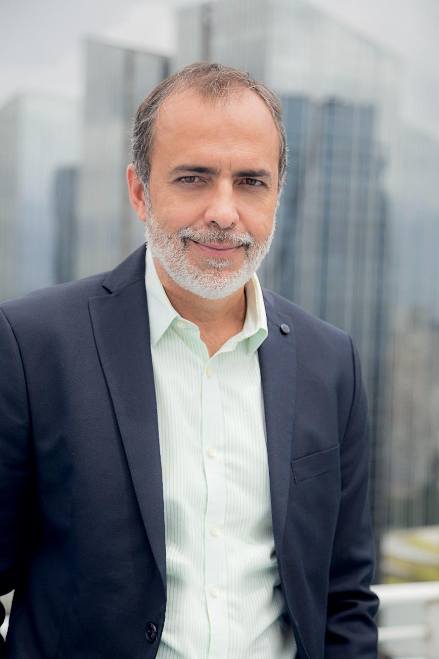 Retrato de um homem de meia-idade com barba grisalha, vestindo um blazer escuro sobre uma camisa verde-clara listrada, sorrindo levemente, com um fundo desfocado de arranha-céus.