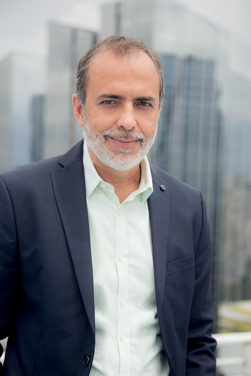 Retrato de um homem de meia-idade com barba grisalha, vestindo um blazer escuro sobre uma camisa verde-clara listrada, sorrindo levemente, com um fundo desfocado de arranha-céus.