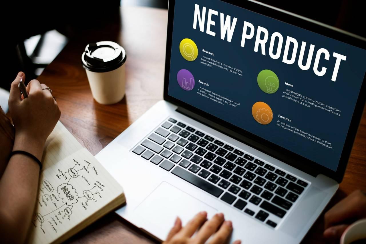 Laptop displaying "New Product" with sections on research, analysis, ideas, and function; a person takes notes nearby.