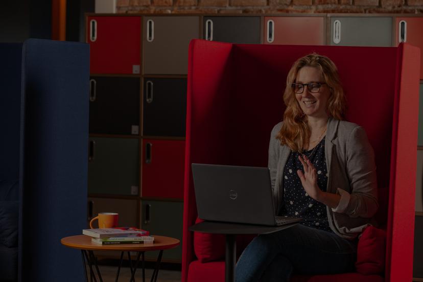 Uma mulher sorridente com óculos senta em uma poltrona vermelha, usando um laptop e acenando para a câmera em uma sala de escritório moderna.