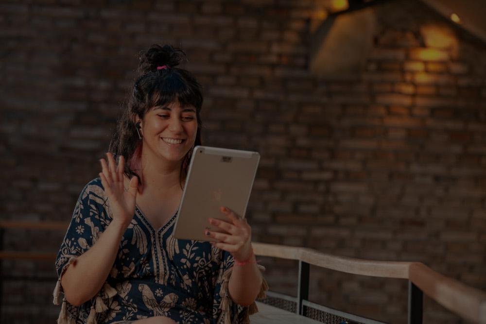 Uma mulher sorridente acena para um tablet enquanto participa de uma videochamada. Ela está sentada em um banco diante de uma parede de tijolos escuros.