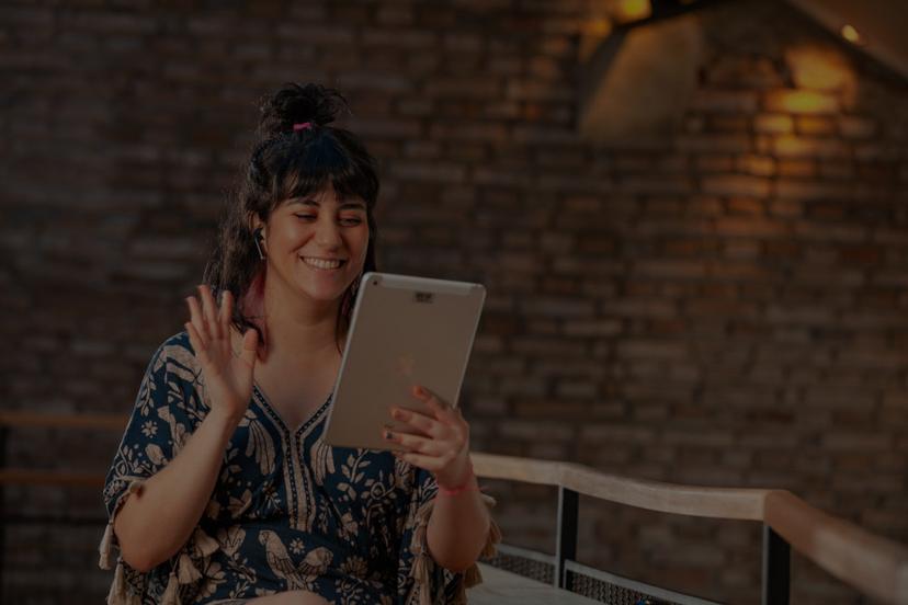 Uma mulher sorridente acena para um tablet enquanto participa de uma videochamada. Ela está sentada em um banco diante de uma parede de tijolos escuros.