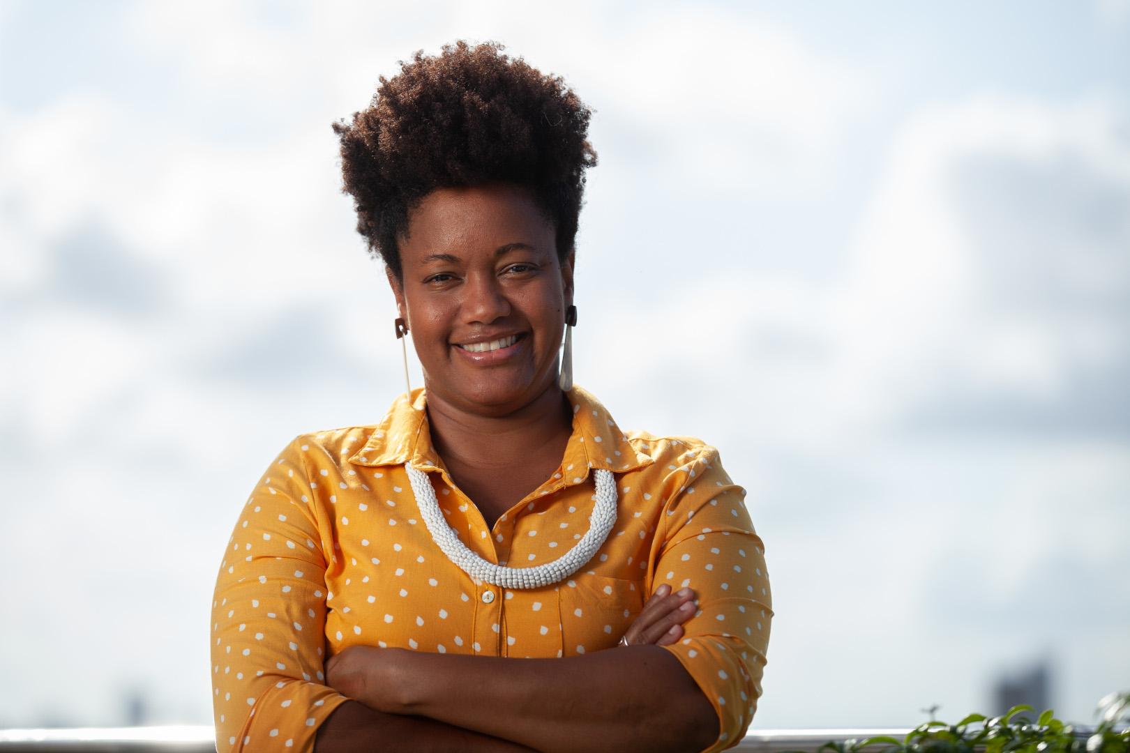 Uma mulher negra sorridente, com cabelo afro, usa uma blusa amarela de bolinhas e um colar branco, com os braços cruzados.
